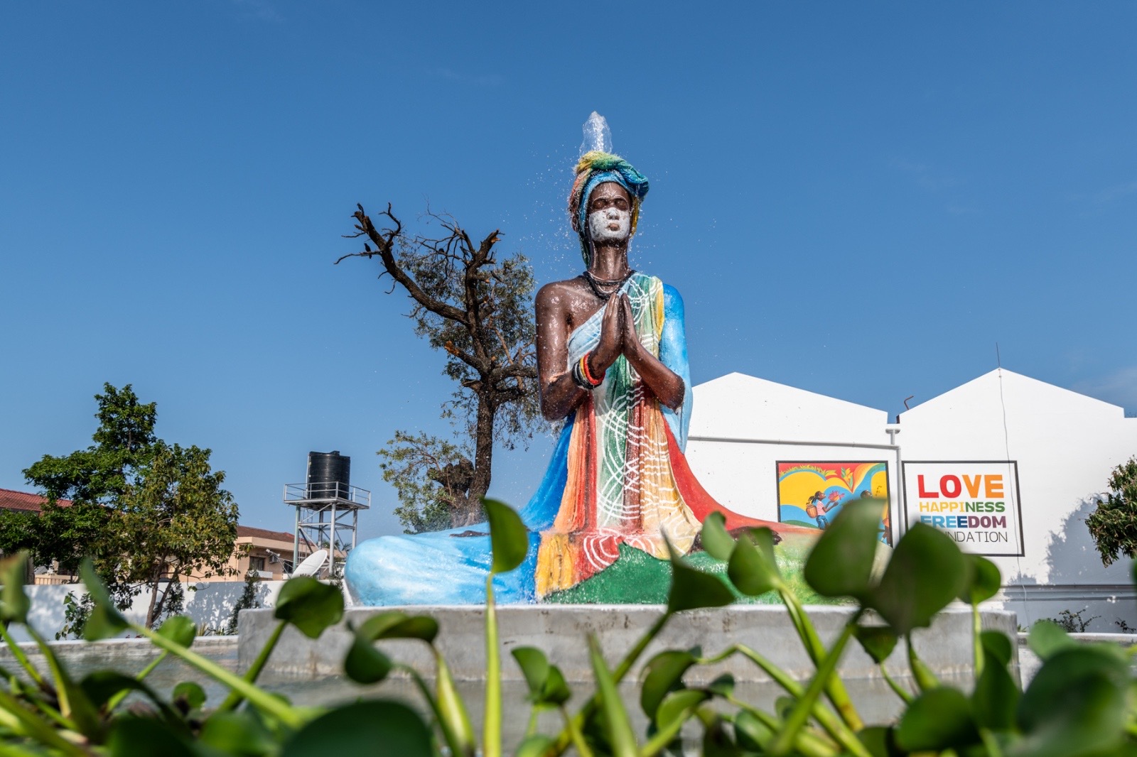 The Love Happiness Freedom school: rainbow-painted fountain sculpture with the Foundation mural and wordmark behind it, daylight.